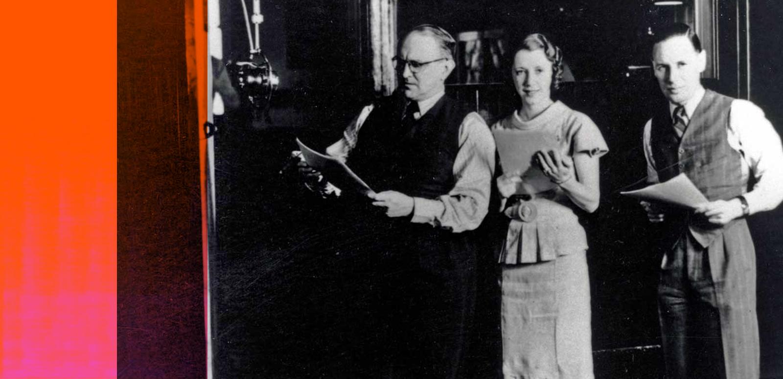 A man and two women in a recording studio standing next to a big radio microphone. They are holding scripts in their hands.