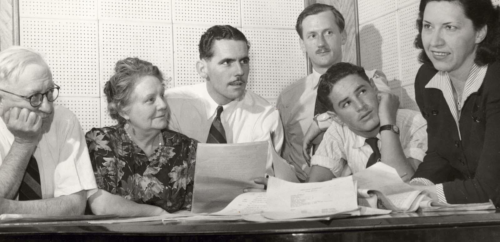 1940s radio series producer Grace Gibson leans on a table with pages of a script while four white men in plain shirts and ties and a woman wearing a floral-patterned dress look on