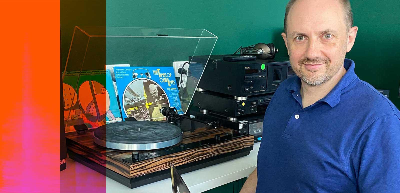 NFSA curator Simon Smith, seated and looking up at the camera. He is sitting at a desk with a turntable and vinyl records on it.