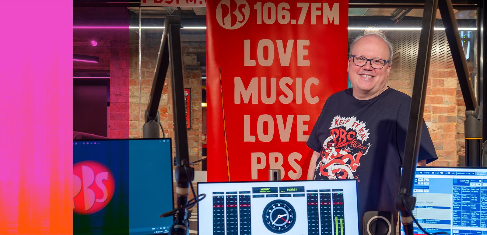 A man stands behind a radio station mixing desk and monitor smiling. He stands in front of a banner that says "106.7FM Love Music Love PBS".