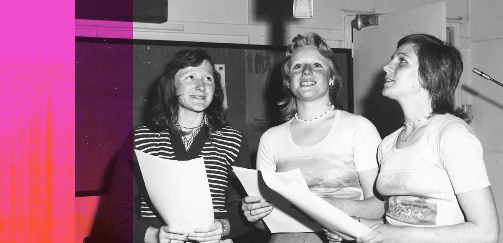 Three women holding scripts in a radio studio are standing around a microphone suspended above them