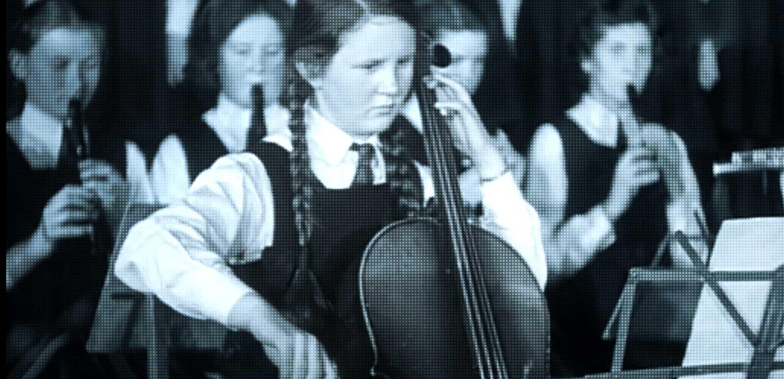 A school girl playing a cello with a group of girls behind her playing recorders.