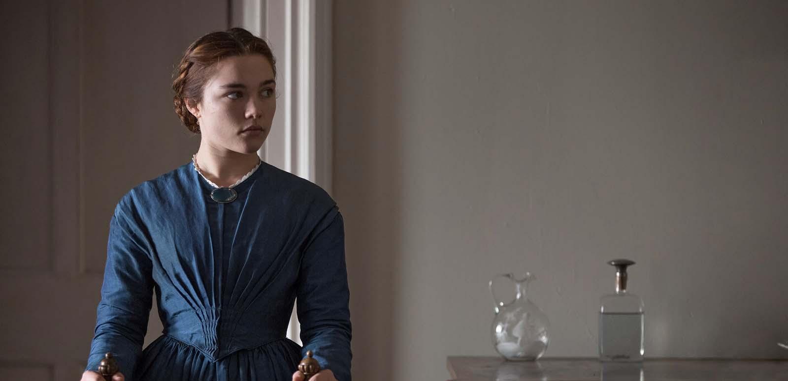 Florence Pugh wearing a blue dress from the 19th century and standing next to a table with a glass bottle and vase on it