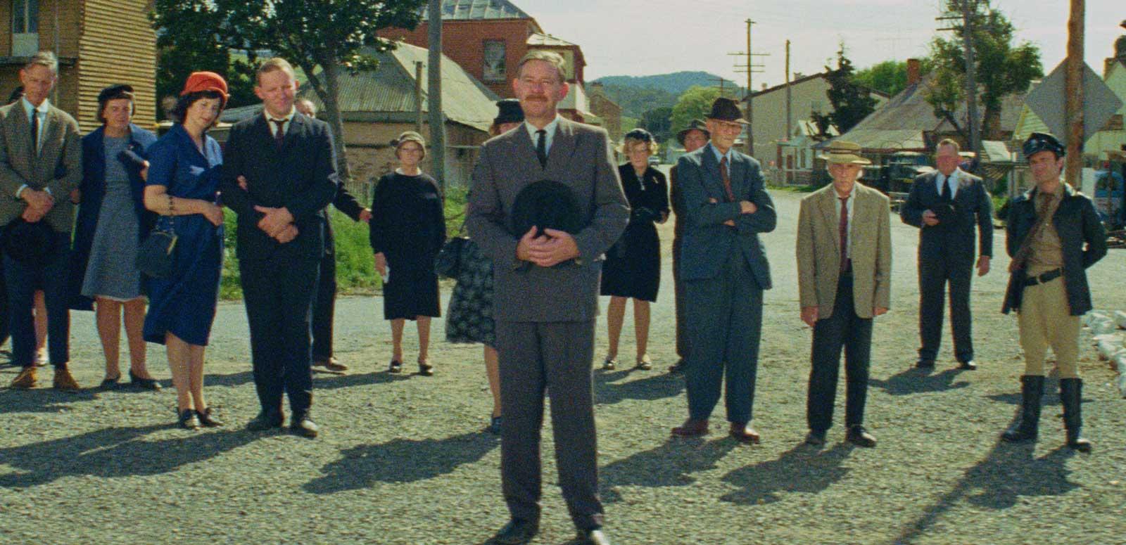 Wide view of a street in a country town. Many of the townspeople are standing in the street looking in the direction of the camera. 
