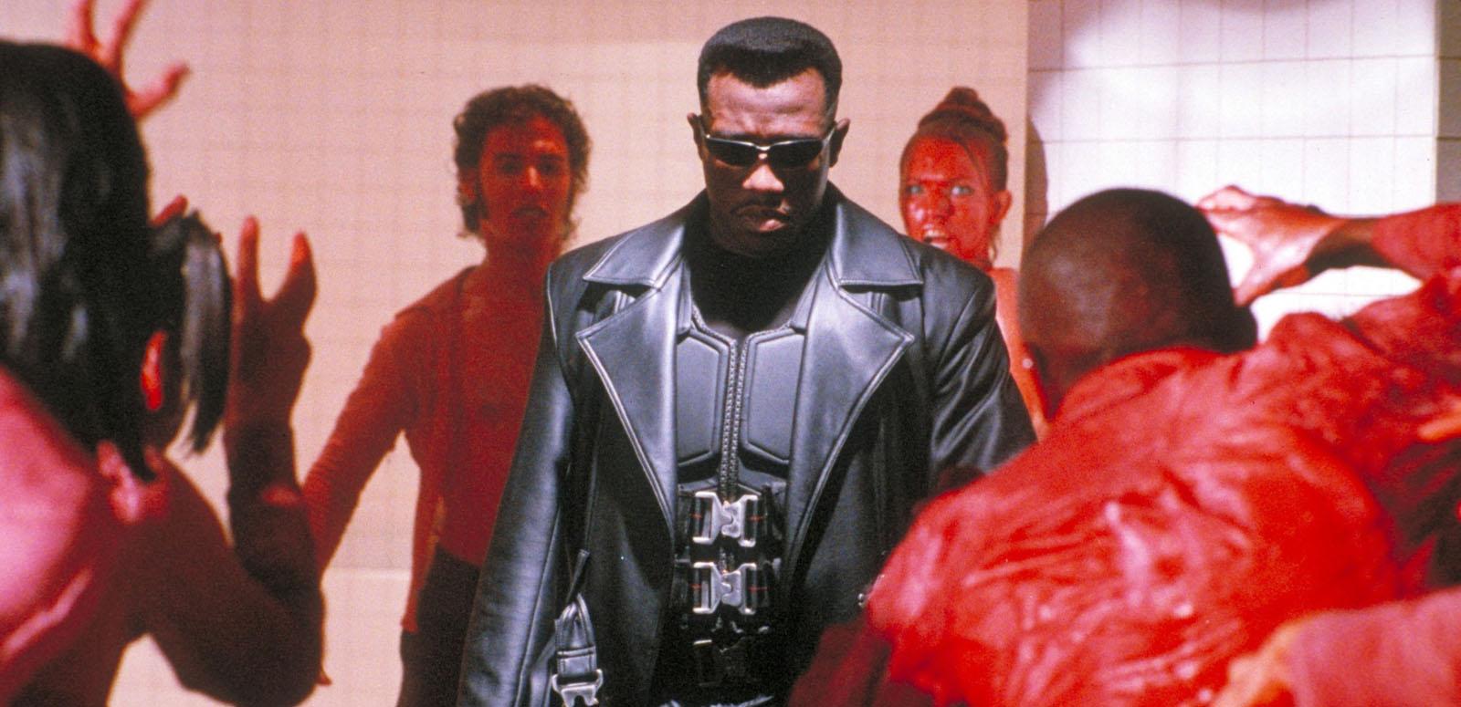Wesley Snipes wearing a black leather trench coat and sunglasses and surrounded by menacing figures with red faces and outfits