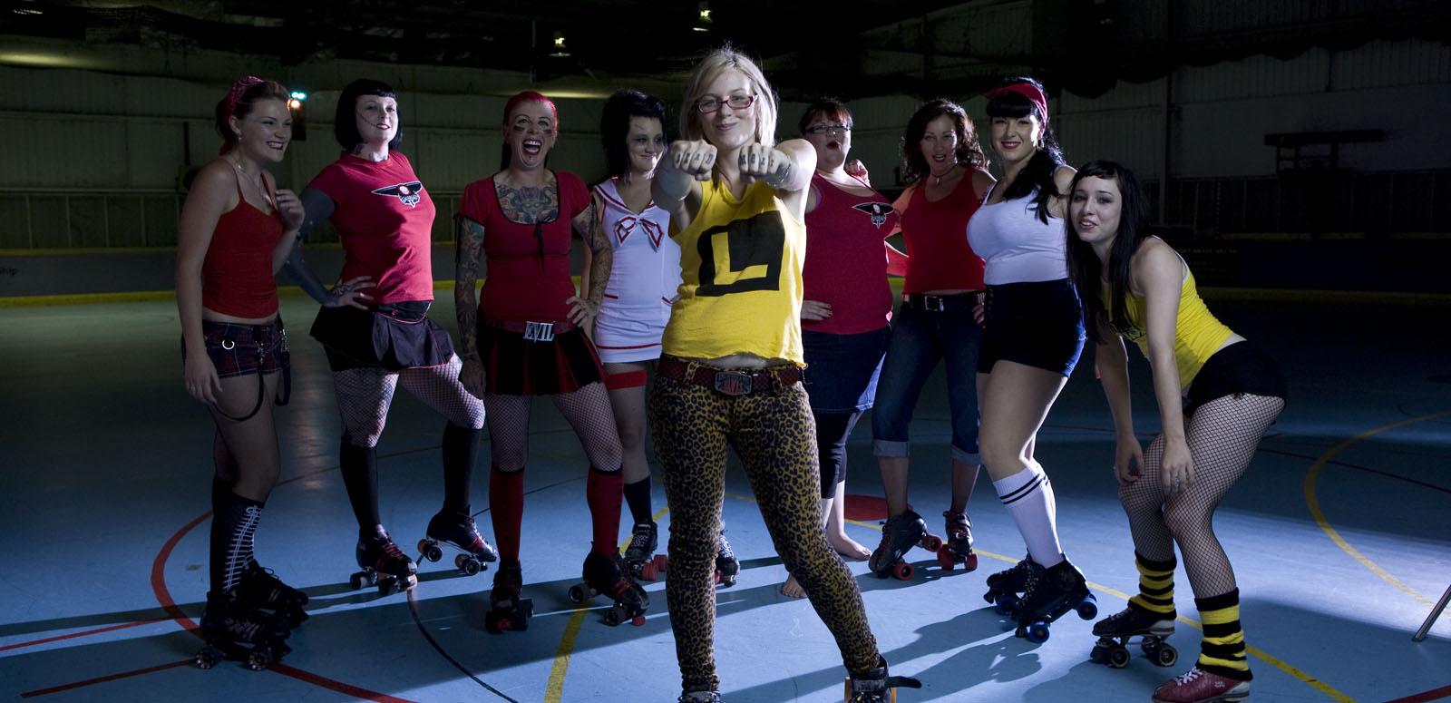 Nine female roller derby players wearing roller skates and standing in a semi-circle, posing. The middle player stands in front of the others and has her arms outstretched and hands bunched into fists.