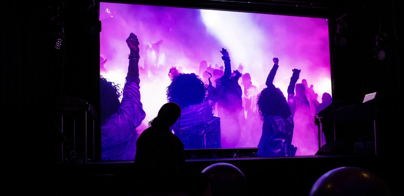 Someone stands in front of a cinema screen projecting three people with their backs to the camera standing in a crowd raising their fists in a swirl of purple smoke