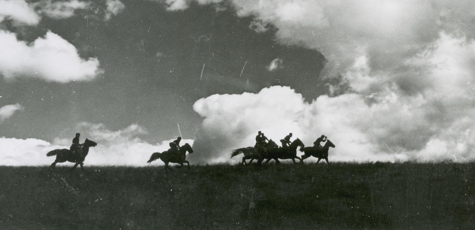 Five horses with riders seen in silhouette riding across a flat landscape beneath a cloudy sky