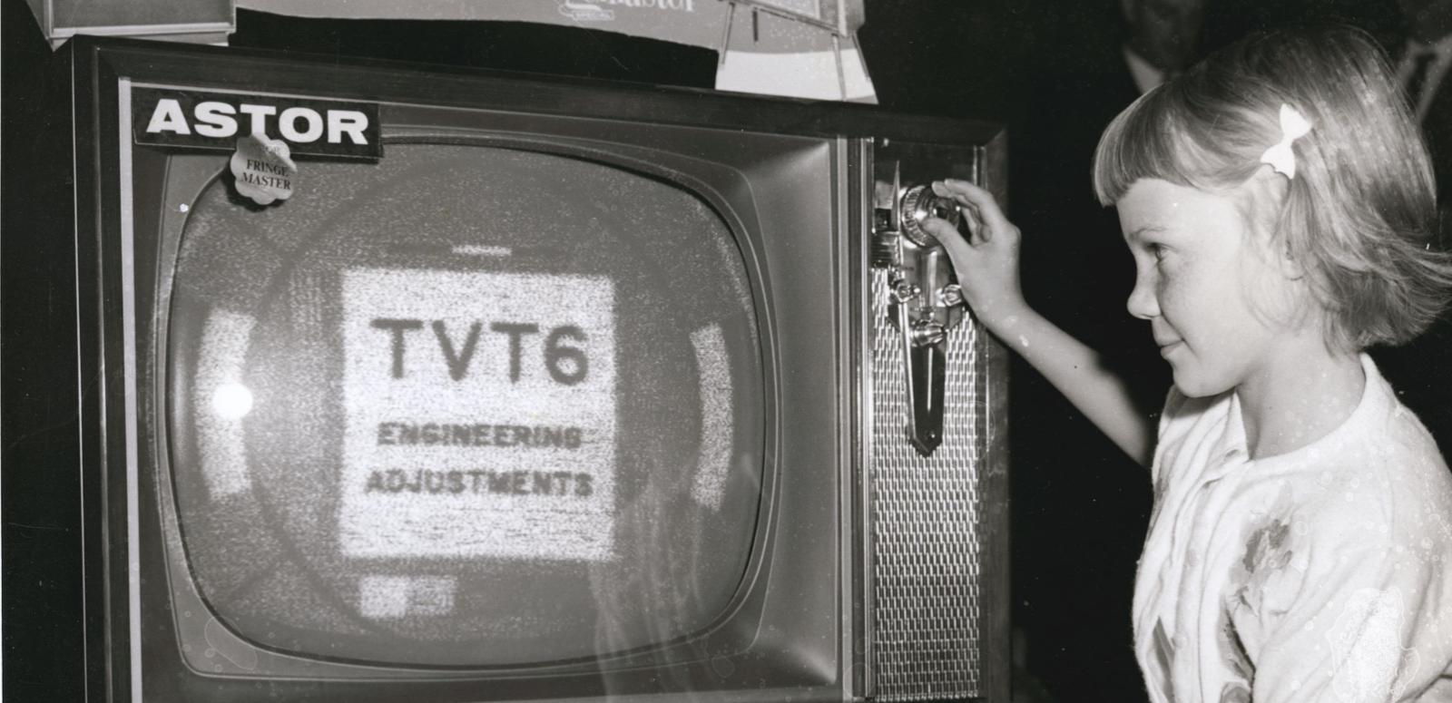 A child adjusts a knob on a large 1960s television set which displays a message "TVT6 engineering adjustments".