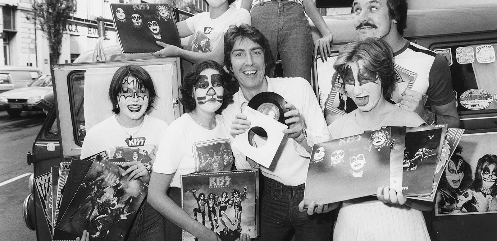 Radio announcer Ron E Sparks surrounded by fans of the band Kiss who have painted faces and are holding up Kiss albums and photos.