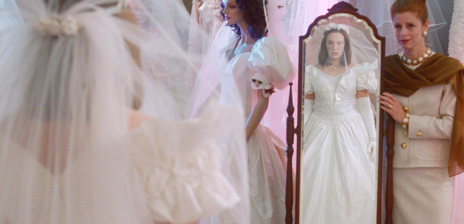 A young woman in a wedding dress looking at herself in a full length mirror while an older woman looks on, in a scene from the film Muriel's Wedding. 