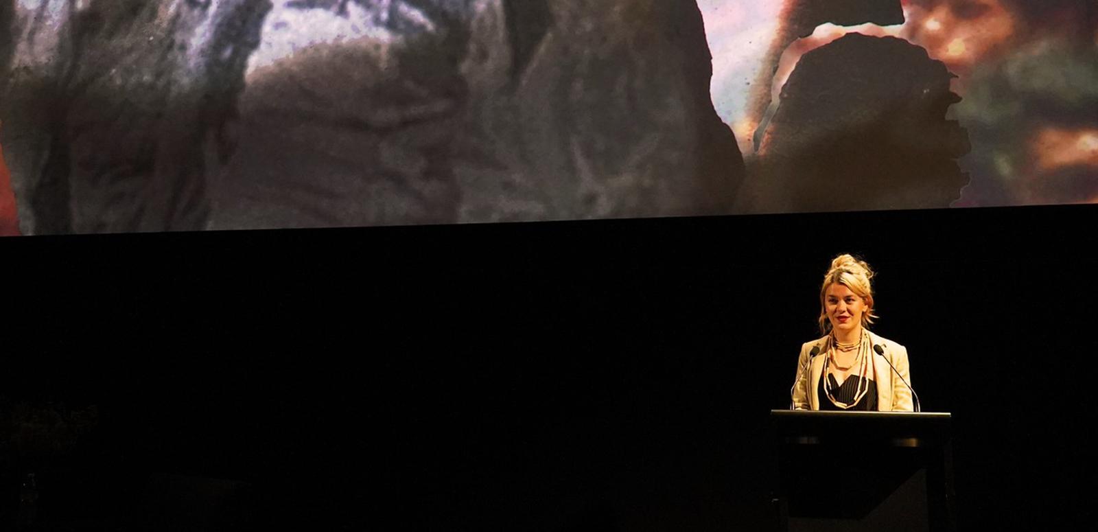Jazz Money standing at a lectern in front of a cinema screen displaying an image from their film WINHANGANHA