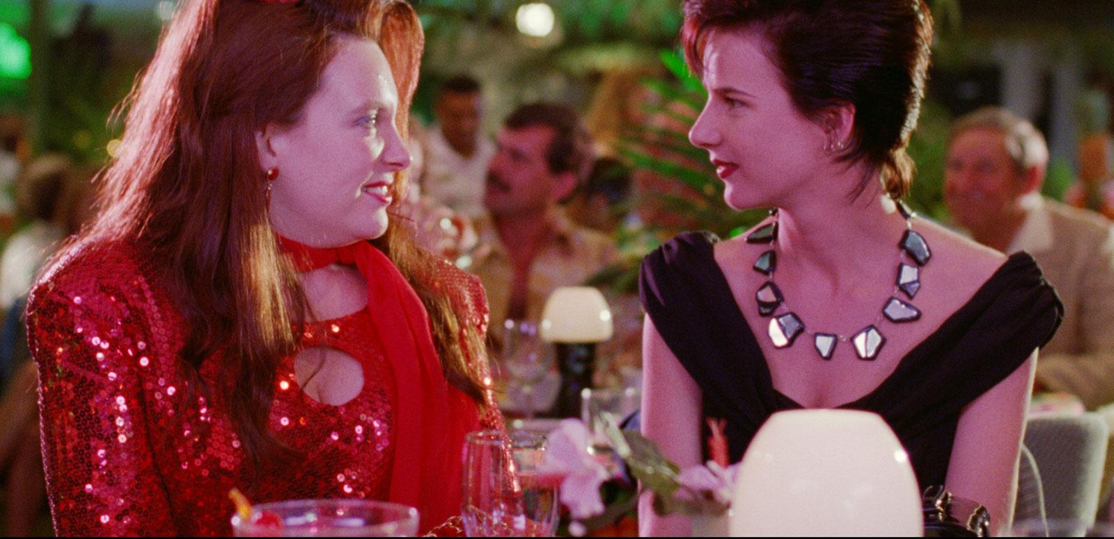 Two women, dressed up for a night out, are sitting at a dining table talking to each other in a scene from Muriel's Wedding.