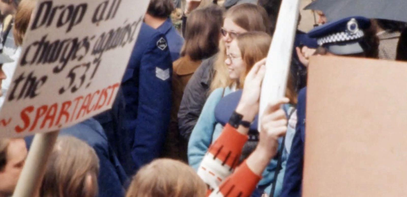 Two women standing in a crowd of protesters and police. A placard near them reads "Drop all charges against the 53".