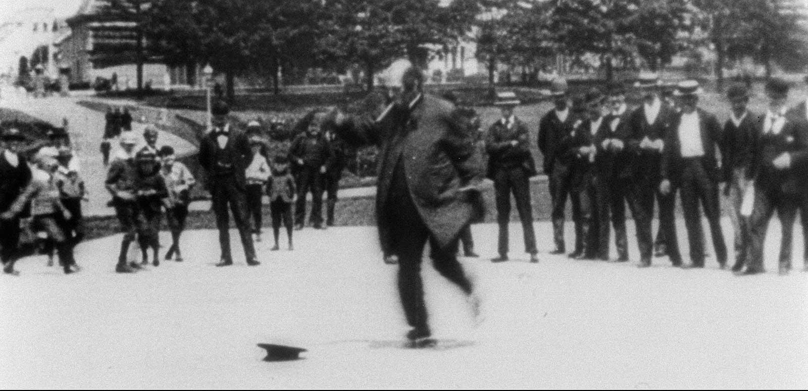 A man skating in a park in front of a crowd of people, circa 1896.