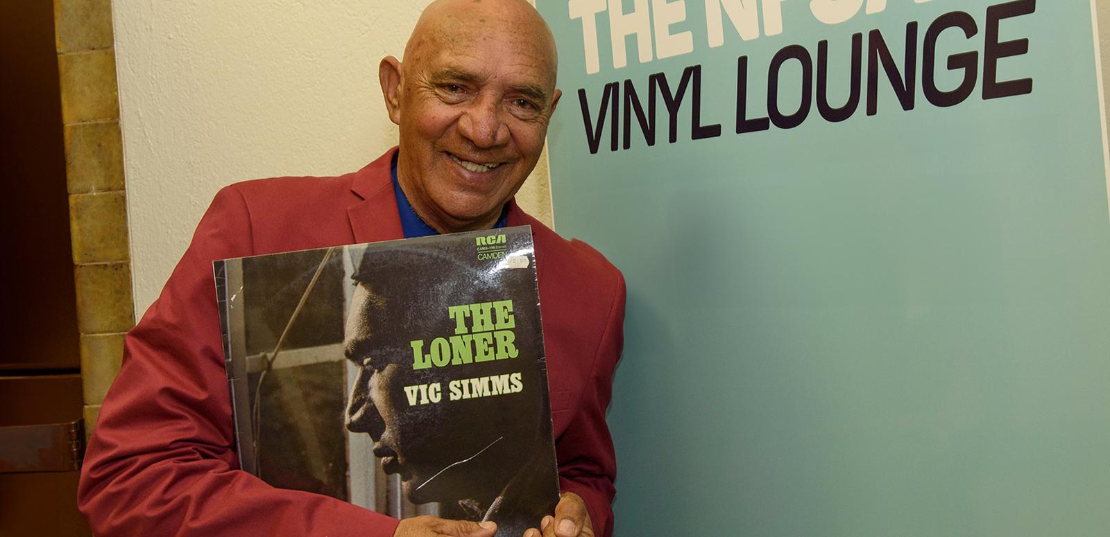 Vic Simms holding up a copy of his vinyl record "The Loner" and standing in front a banner advertising the NFSA's Vinyl Lounge