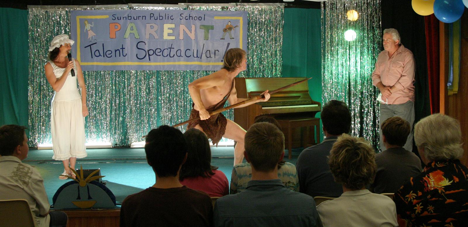 On stage at a school parents' talent show, a woman in a frilly hat and old-fashioned nightgown holds the microphone while a man in a loincloth holds a spear and lunges towards the compere standing in the wings.