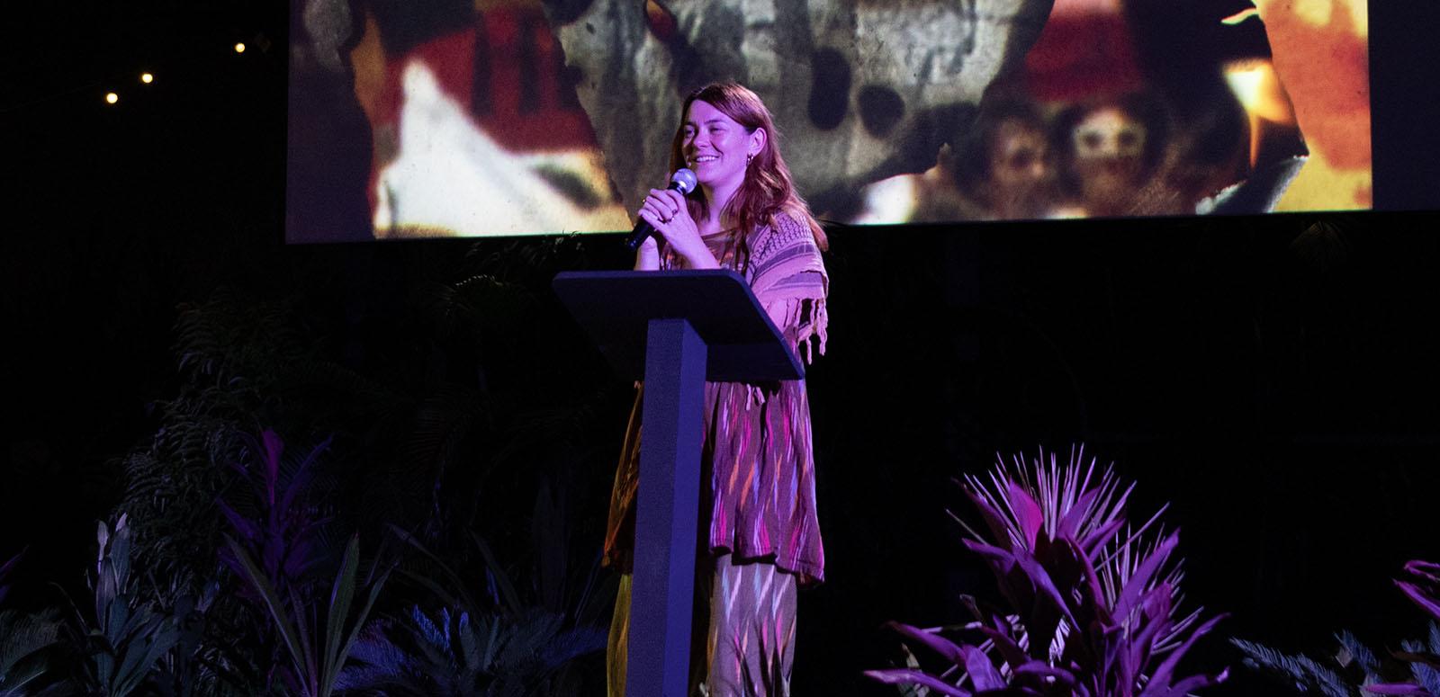 Jazz Money standing at a lectern in front of a cinema screen displaying an image from their film WINHANGANHA