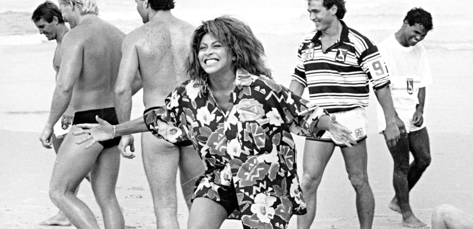 Tina Turner laughing on a beach, surrounded by male rugby league players in swimming costumes.