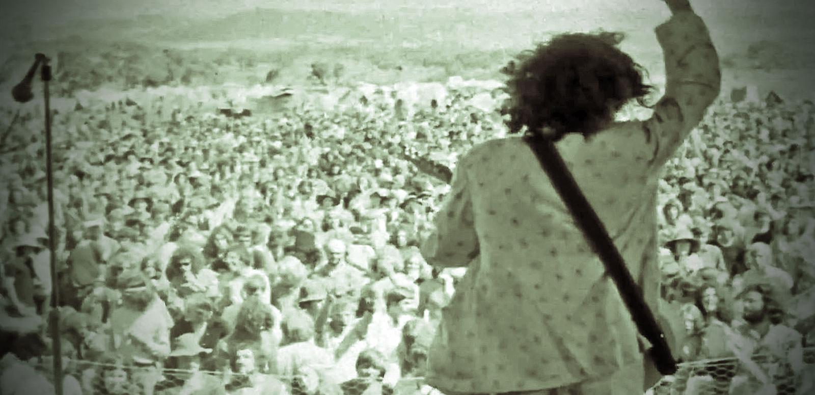 Eagle Rock hero image Ross Wilson playing guitar on an outdoor stage with his back to camera, facing a large crowd with his right arm raised, from a screenshot of the film clip for the song Eagle Rock.