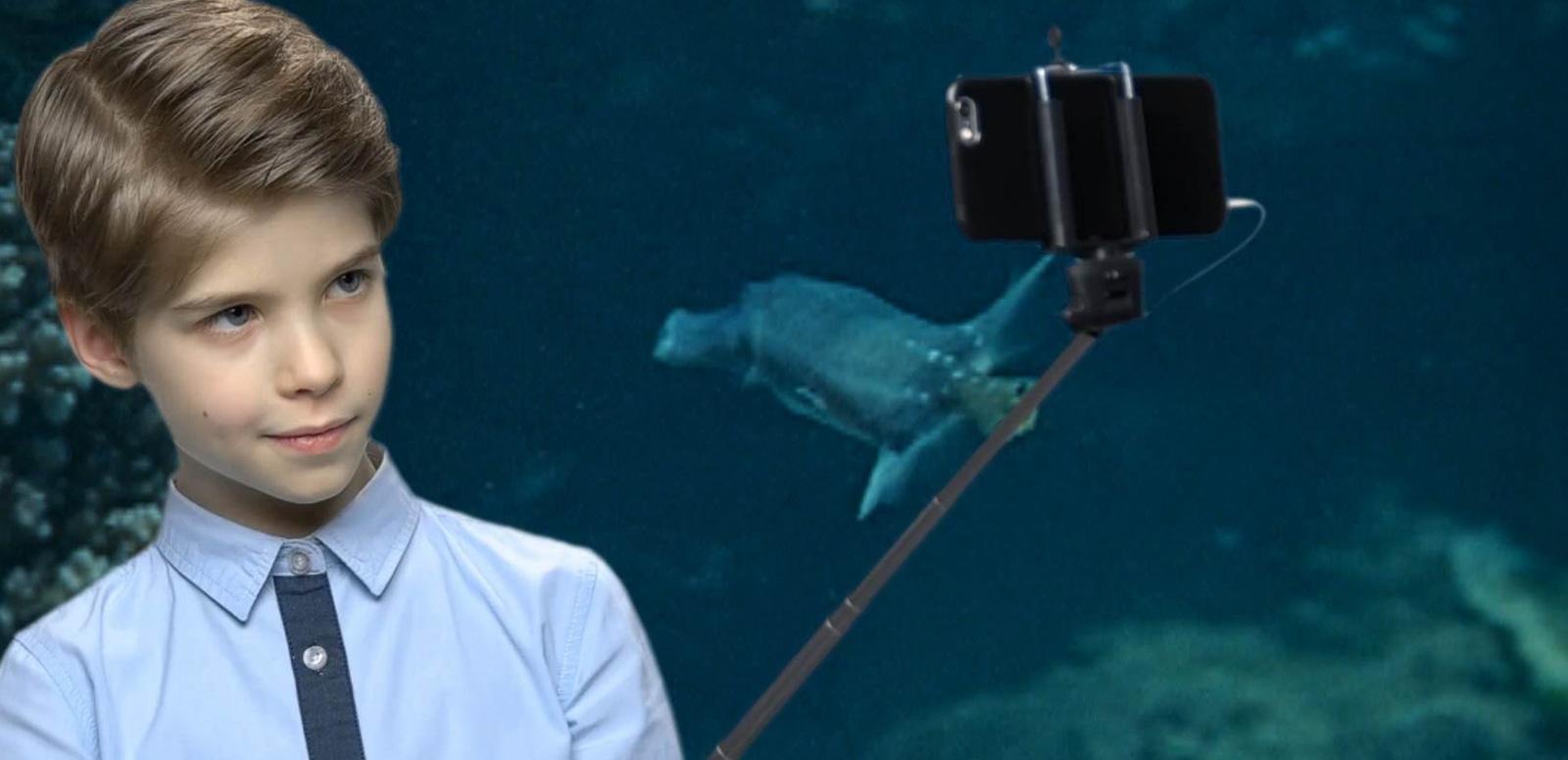 A boy holds a phone on a selfie stick against the backdrop of a video screen showing an underwater scene. A boy holds a phone on a selfie stick against the backdrop of a video screen showing an underwater scene.