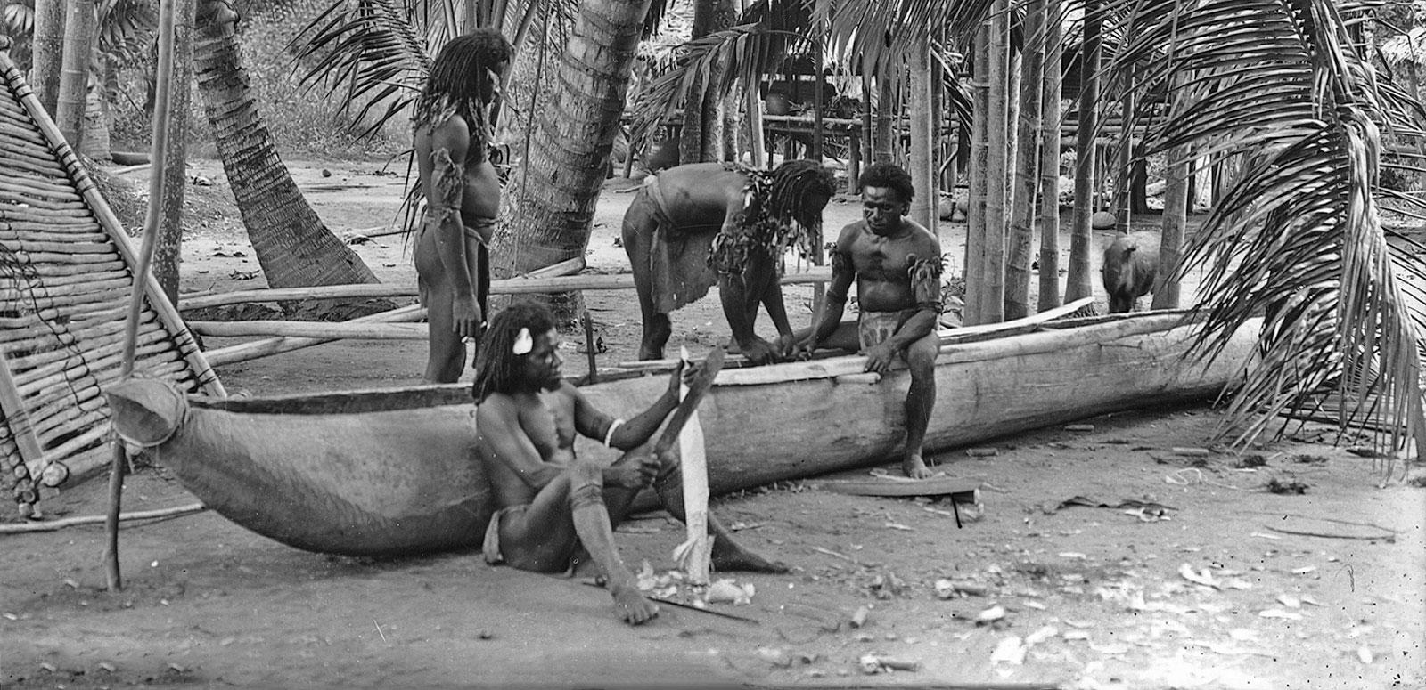 A group of Torres Strait Islander men building a canoe by the water, surrounded by palm trees in the early 1920s.