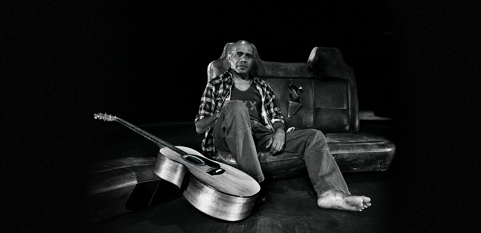 Archie Roach - Bangarra Archie Roach rests against an old car seat in a studio with his guitar resting nearby.