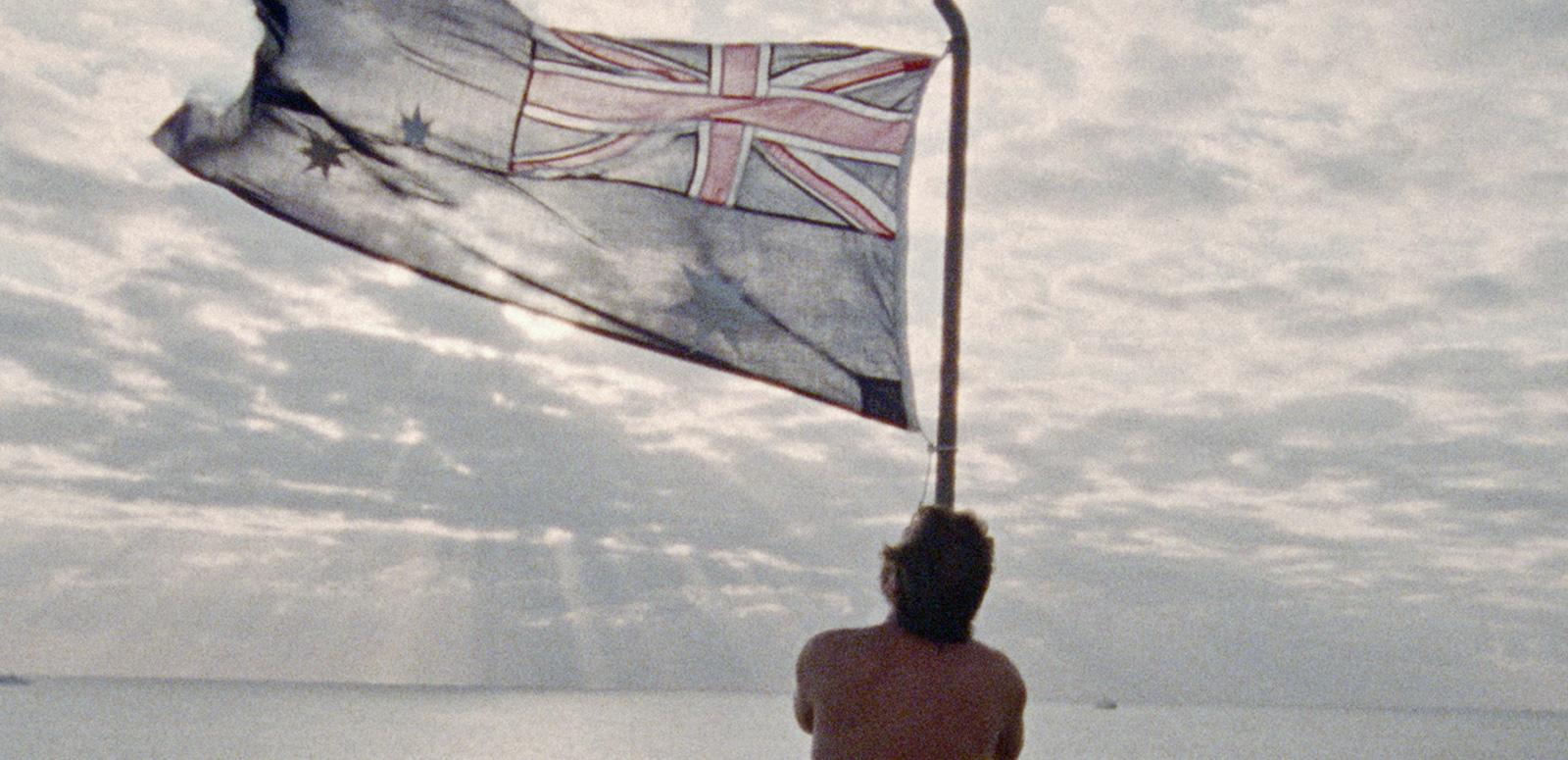 Man waving an Australian flag, set against a pale blue, cloudy sky.