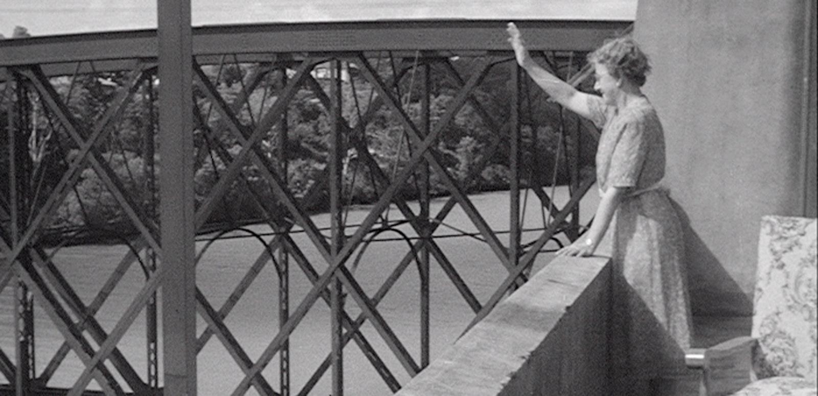 Brisbane time capsule article hero A woman is waving at someone we can't see. She is standing on a balcony with girders of a bridge nearby.