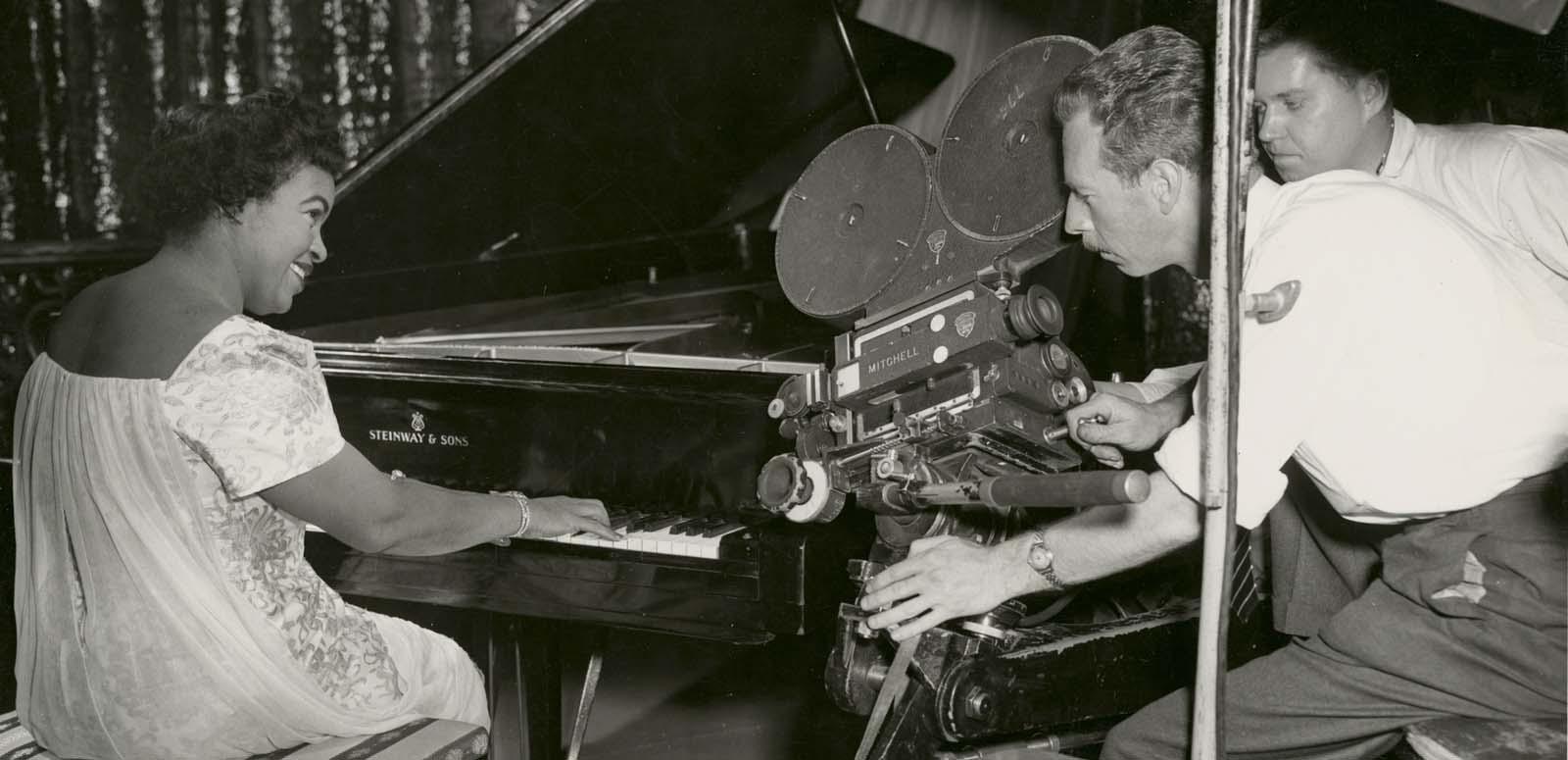 Winifred Atwell playing the piano and smiling while two men film her with a 35mm film camera next to the piano Winifred Atwell playing the piano and smiling while two men film her with a 35mm film camera next to the piano