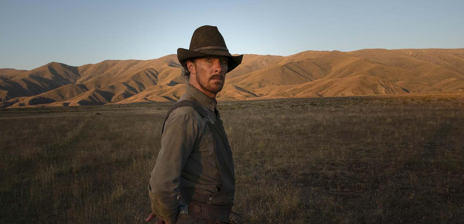 Benedict Cumberbatch dressed in weathered rancher's hat and overalls stands upright on his Montana farm, staring at the camera