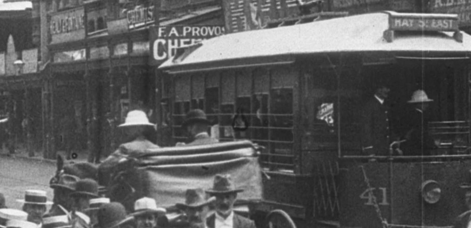 Street Scenes in Perth A tram goes past on a busy street in 1907. A horse and carriage is also going past. Men all wear hats. Shop hoardings promote businesses.