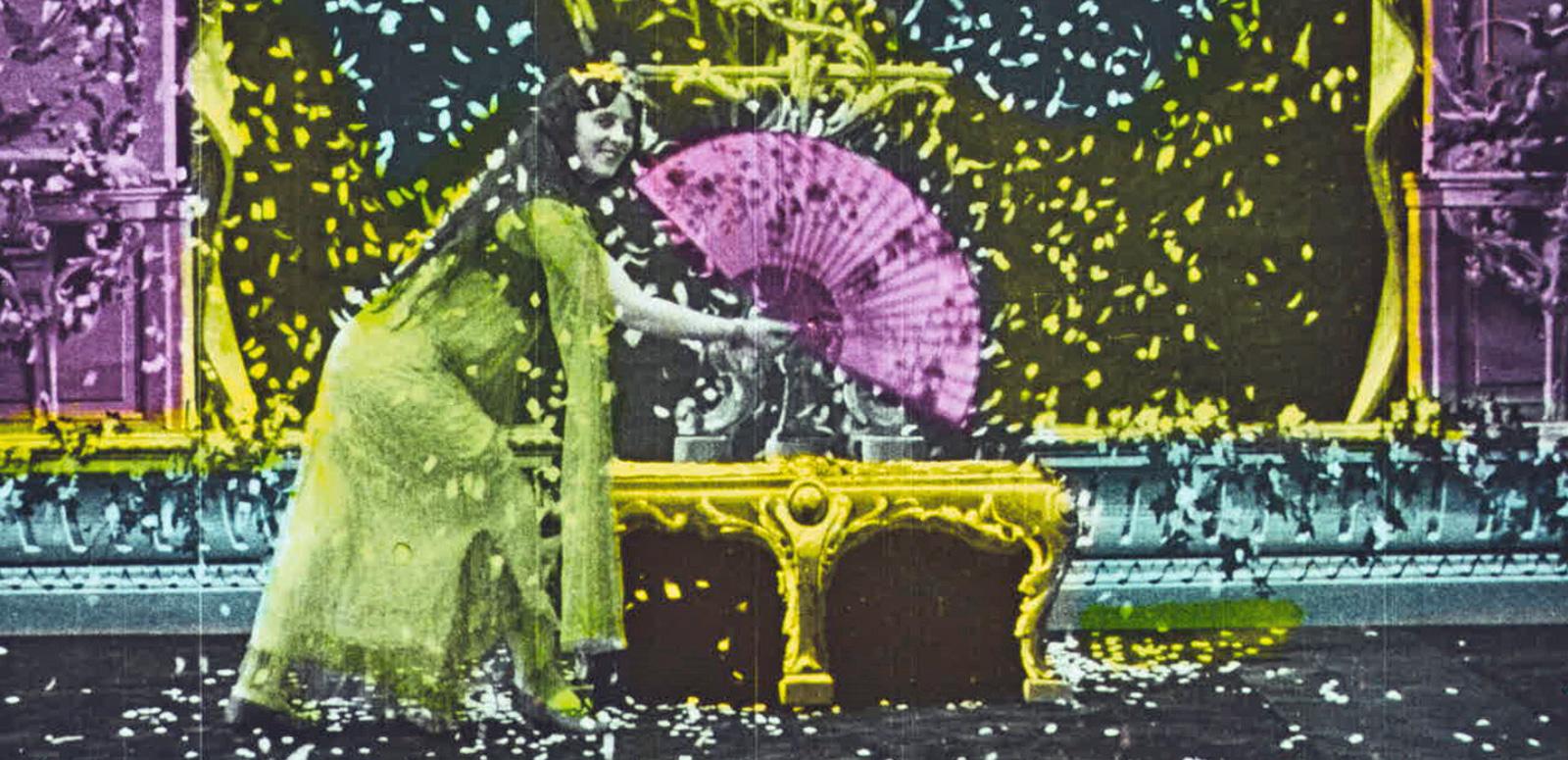 La Fee Aux Pigeons Still from a colourised film showing a woman bent down near a table holding a large fan.