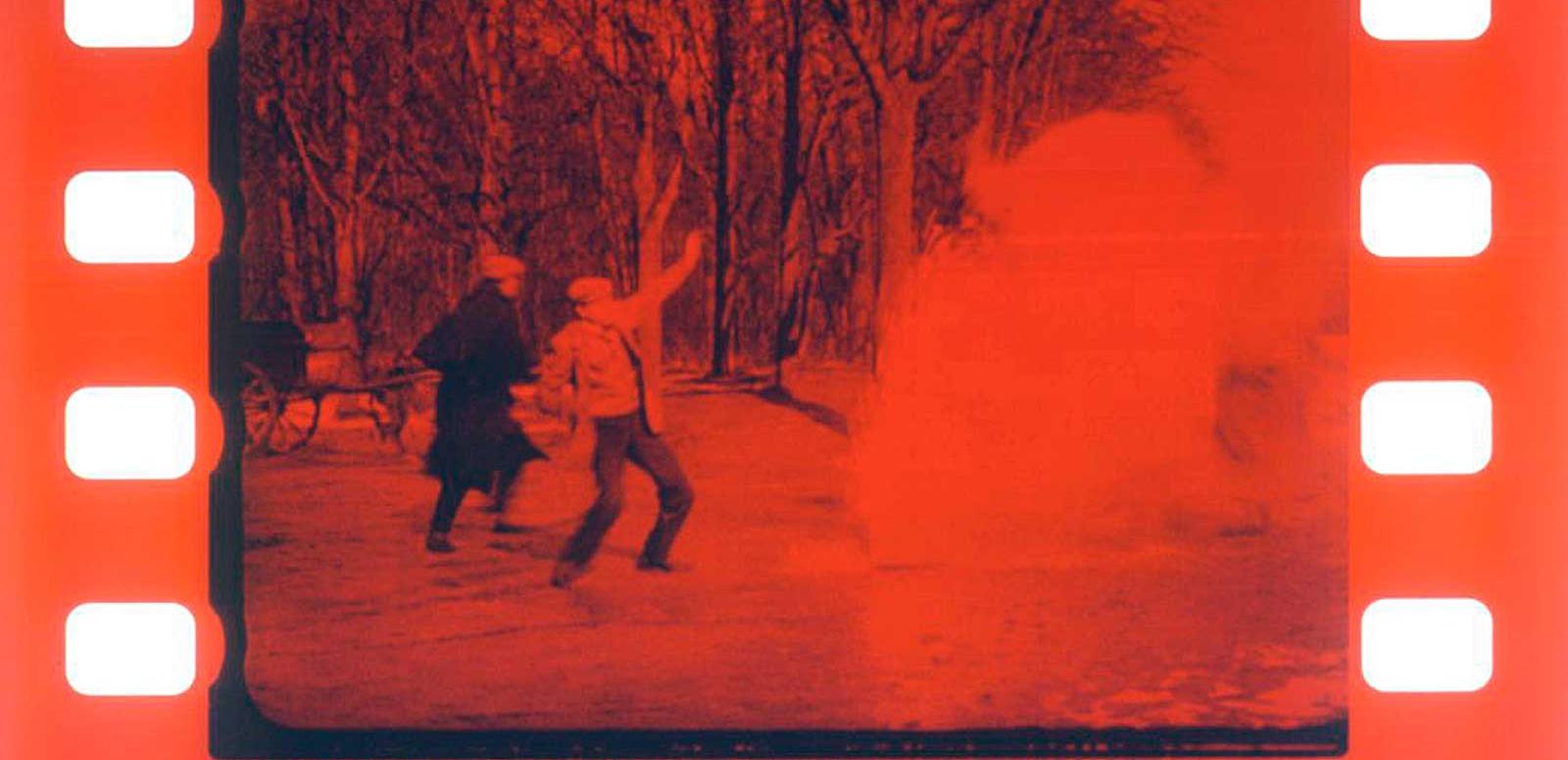Frame of early silent film that has been tinted red showing two men in a forest pointing to the sky