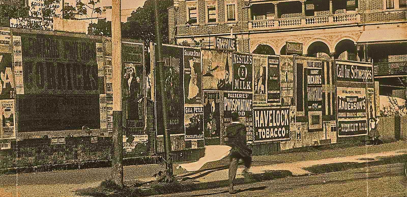 A scene from the early silent film Bashful Mr Brown shows a man walking down a street next to a fence lined with posters and advertisements.