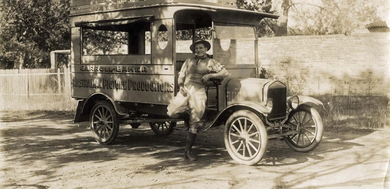 Actor and filmmaker Snowy Baker leaning against a car with 'Carroll-Baker Australian Picture Productions' written on the side
