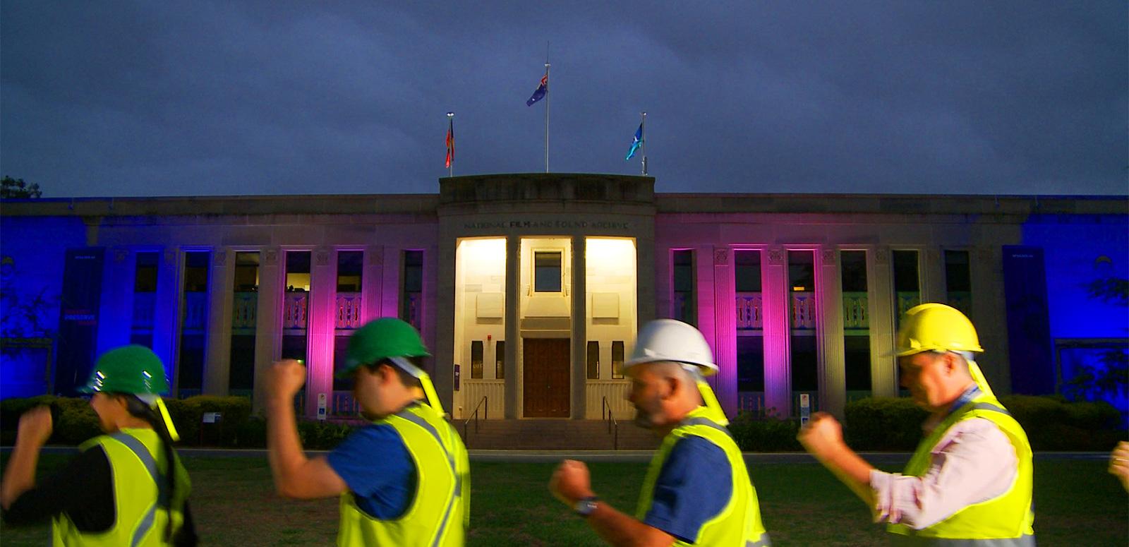 Dance Off_1600x775 Four people wearing protection helmets and hi viz shirst march in line in front of the NFSA building at night.