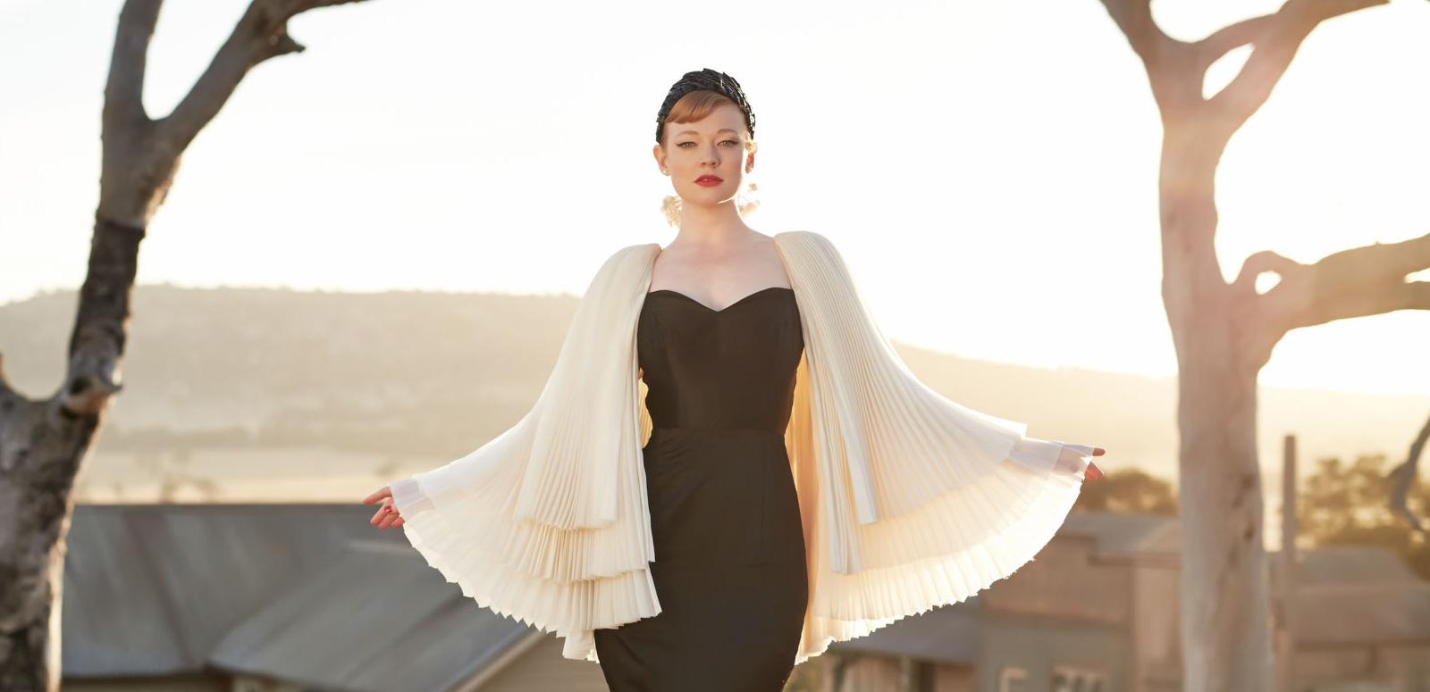 Saran Snook in black dress with white cape stands in front of an outback scene at sunset