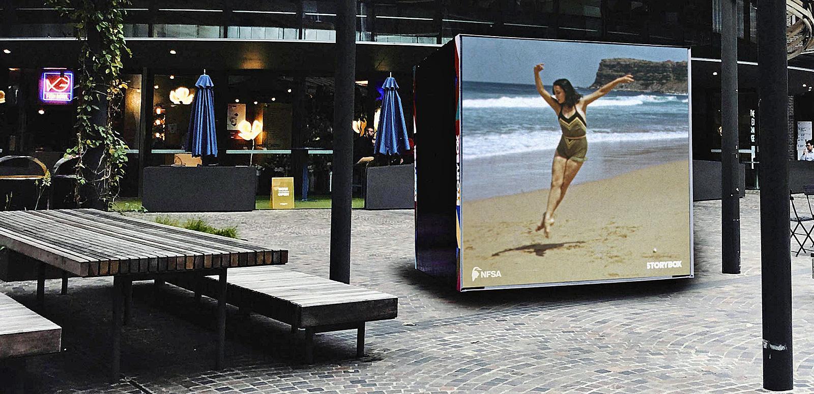 An open area in the middle of town with picnic tables and modern architecture surrounding a large square screen playing a film.