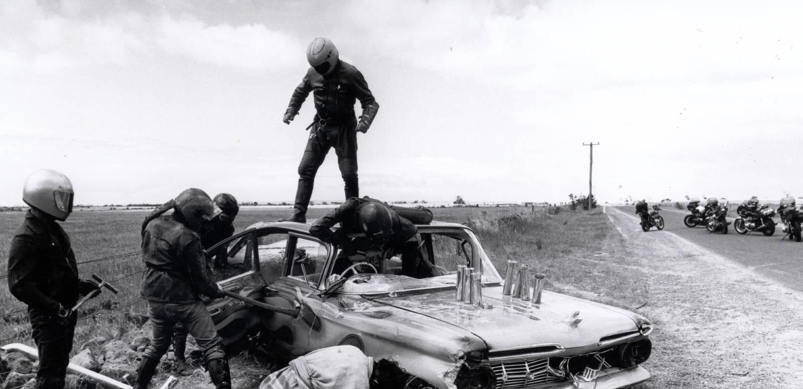 Members of a motorcycle gang smash up a car by the side of a highway
