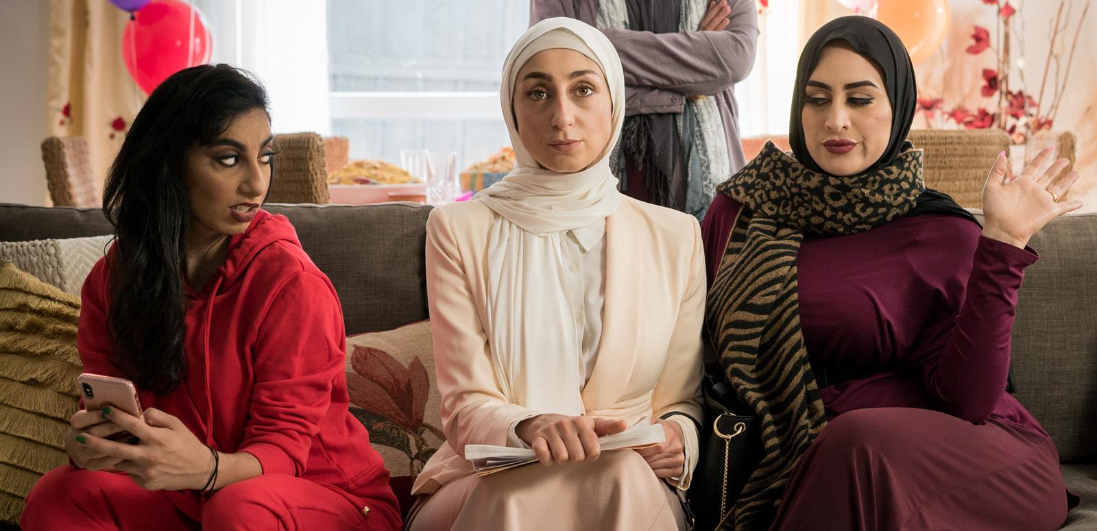 Three women sitting on a couch. One is looking sideways with an expression of annoyance at the other two women who are both looking straight ahead.
