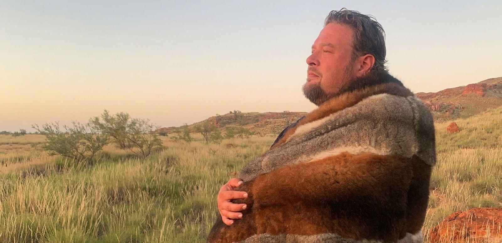 A man standing in profile pictured from the waist up wrapped in a traditional animal skin blanket.