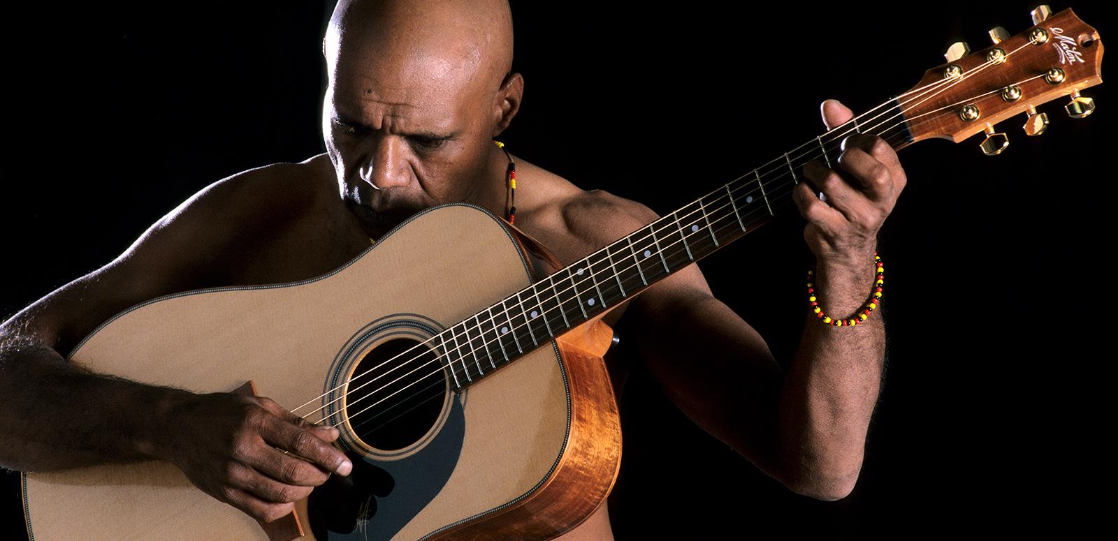 An Aboriginal man holding an acoustic guitar with his head bowed.