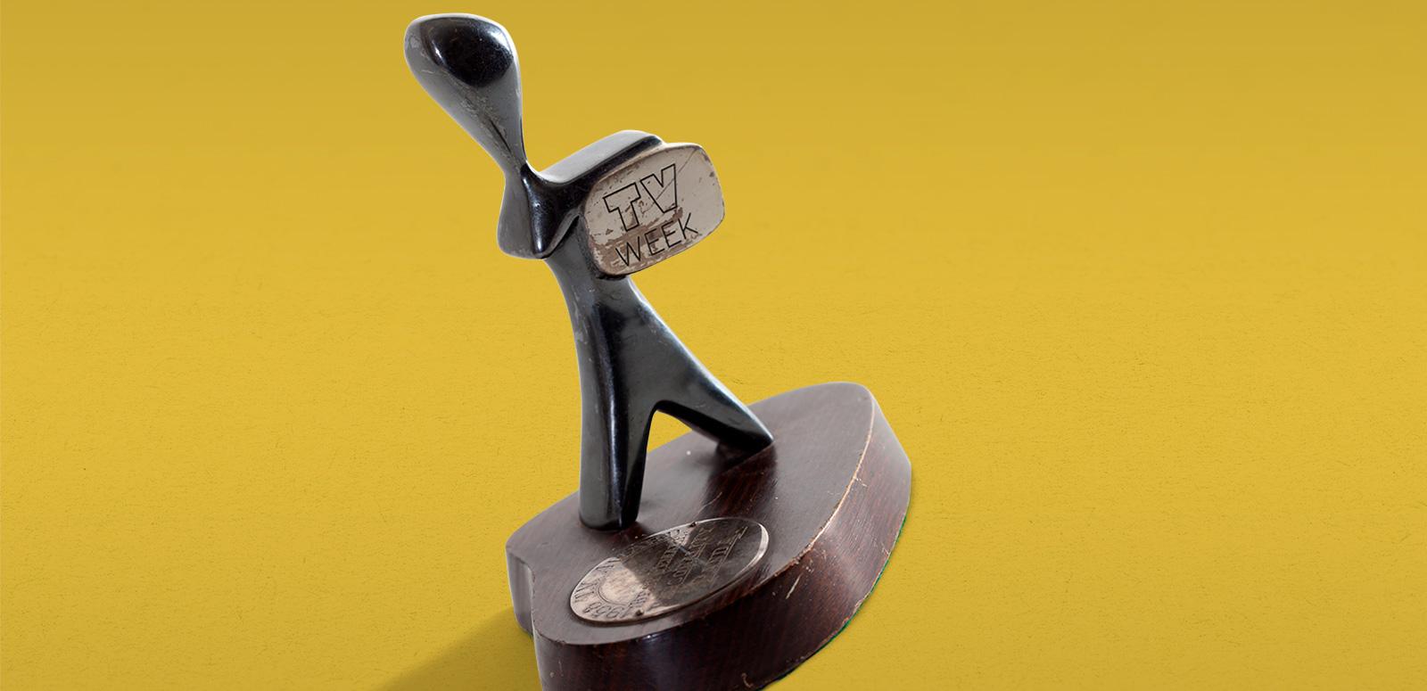 A TV Week Logie Award, made up of a small silver statuette in the shape of a man holding a television. The statuette sits on a round wooden base with a metal disc with words engraved on it. 