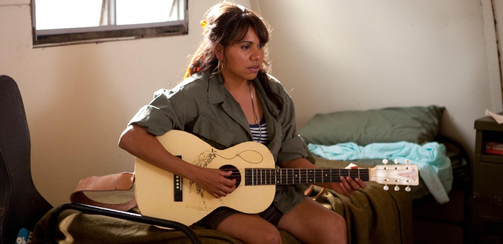 A woman sitting on a bed playing an acoustic guitar.