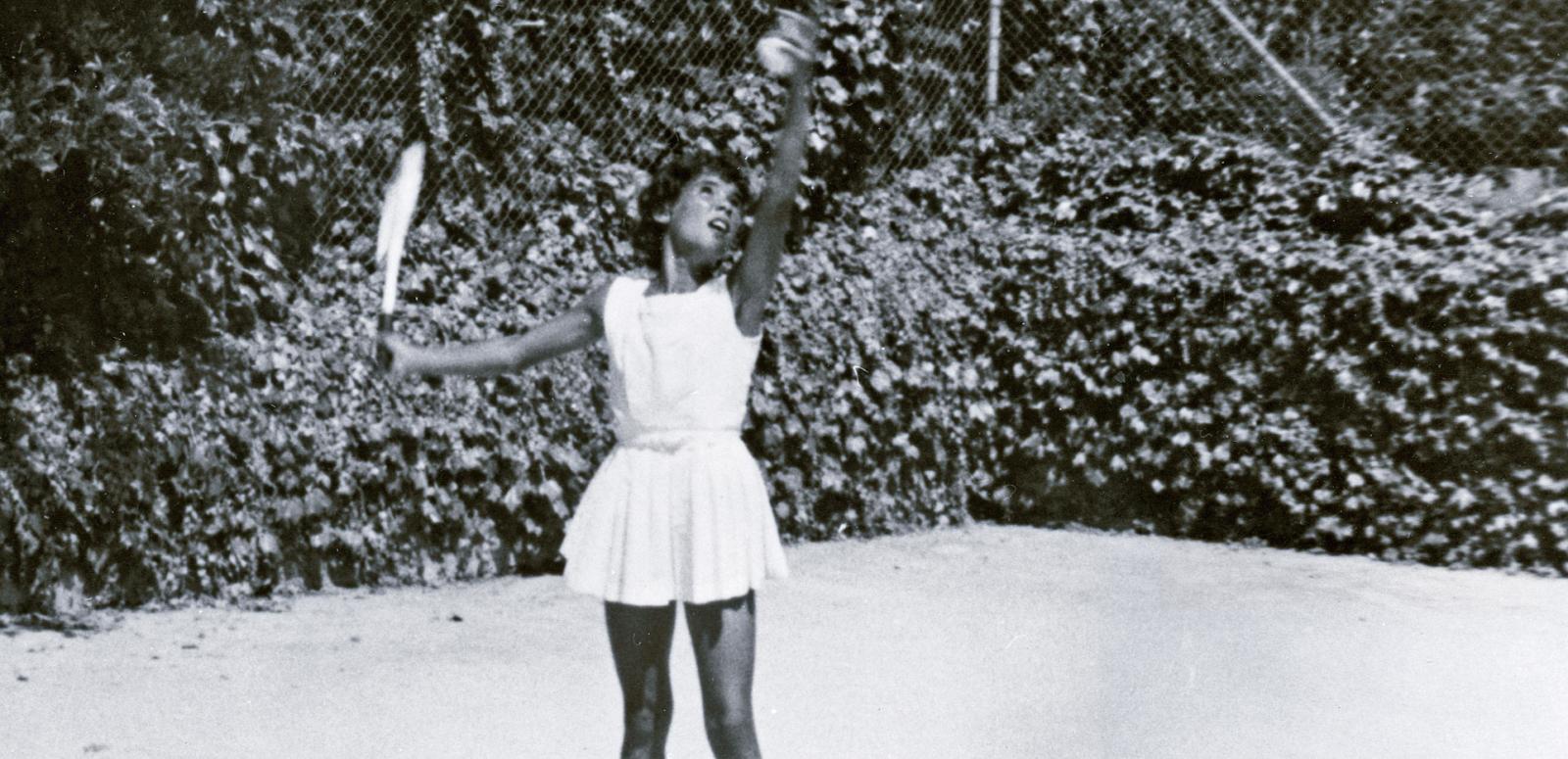 A young Aboriginal woman in a white tennis dress serving a tennis ball. 