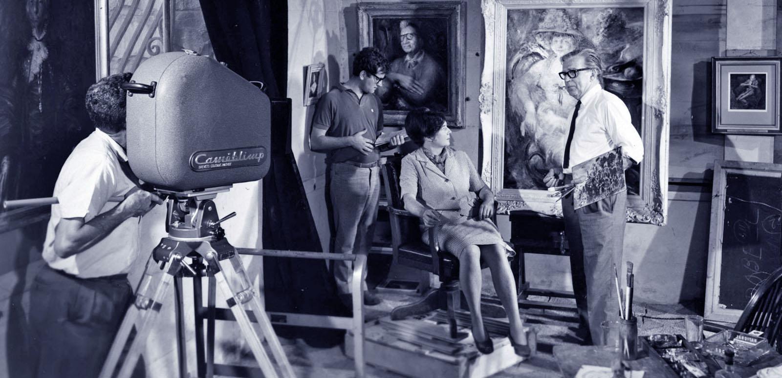 Blue-tinted black and white still of a man is operating a large film camera. He is filming artists Margaret Olley and William Dobell. There are two portraits painted by Dobell in the background as well as another member of the film crew. Blue-tinted black and white still of a man is operating a large film camera. He is filming artists Margaret Olley and William Dobell. There are two portraits painted by Dobell in the background as well as another member of the film crew.