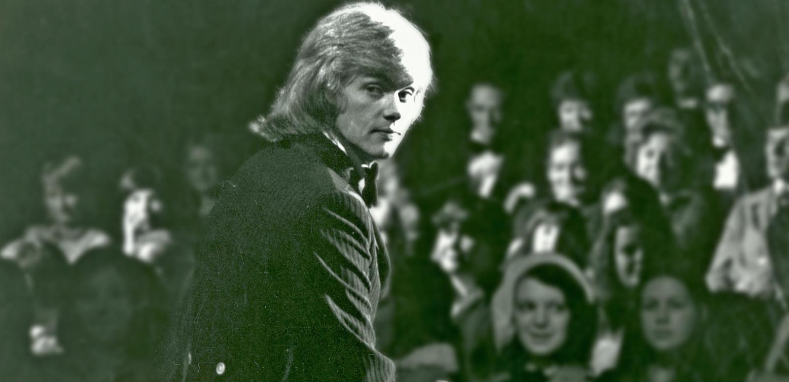 John Farnham sitting on stage in front of an audience, looking back over his right shoulder at the camer.