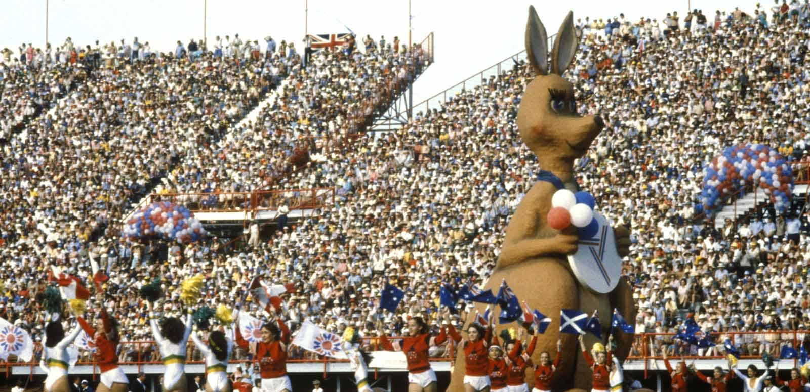 A packed stadium of people witnessing the opening ceremony of the Commonwealth Games. Matilda the huge mascot of the games is on the field along with lots of school children.