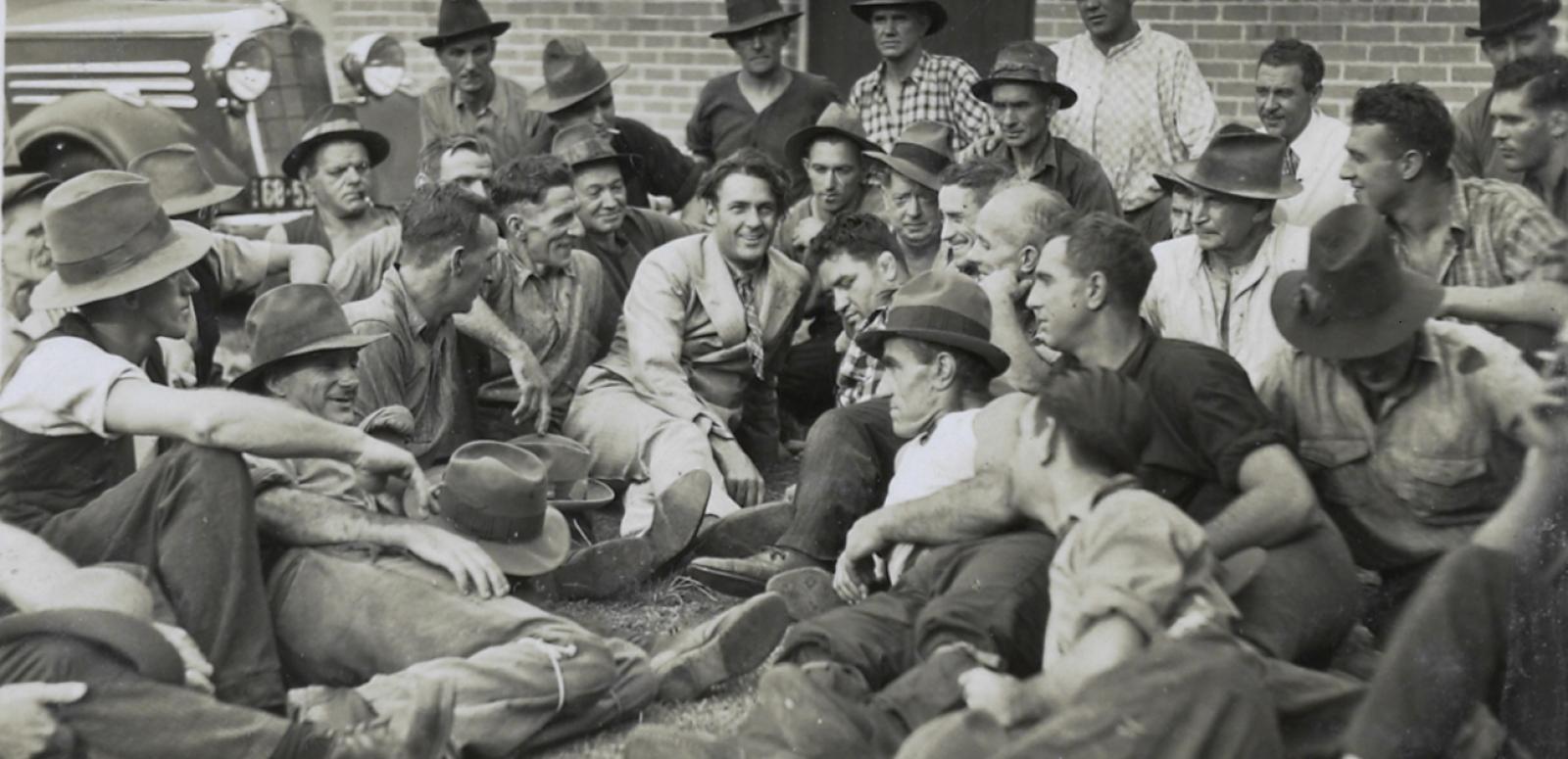 Actor Charles Farrell reclines on the ground surrounded by a crowd of men. Actor Charles Farrell reclines on the ground surrounded by a crowd of men.